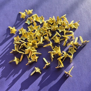 Op de afbeelding: Een grote stapel gouden stervormige metalen bevestigingen. De bevestigingen hebben een puntig uiteinde en een platte, stervormige kop.