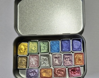 May include: An open, silver-colored metal tin containing a set of 16 watercolor paints. The paints are arranged in small, rectangular pans and display a variety of metallic colors, including gold, green, blue, and purple. The tin is on a white surface.