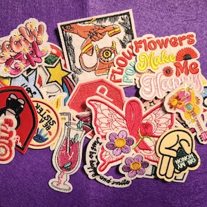 以下が含まれることがあります： 虹、花、蝶、ギター、そして「Flowers Make Me Happy」というテキストなど、さまざまなデザインの刺繍パッチのコレクション。 パッチはさまざまな色と形をしています。