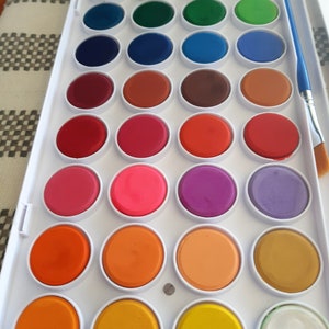May include: A white plastic watercolor paint set with 24 round paint pans. The pans are arranged in four rows of six. The paint colors include red, orange, yellow, green, blue, purple, pink, brown, and black. A blue paintbrush is included.