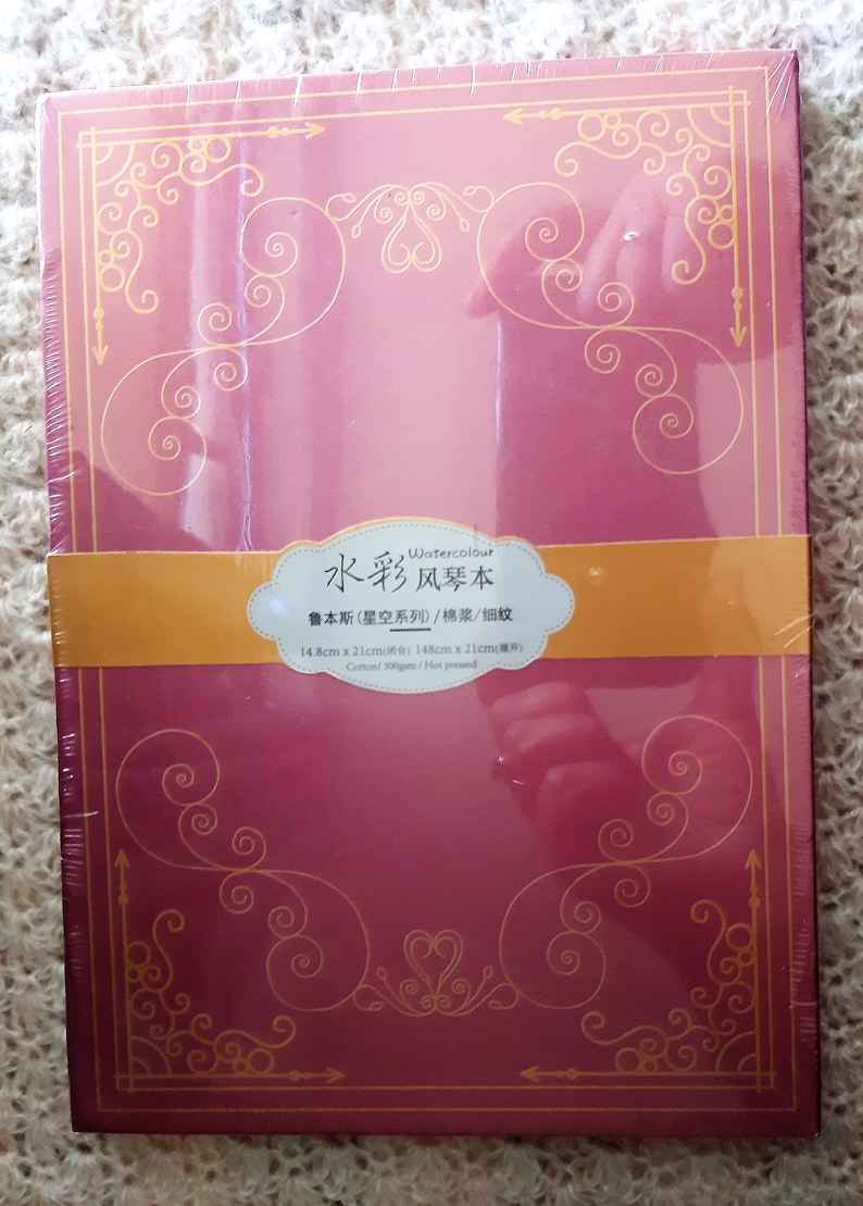 K&ouml;nnte beinhalten: Ein rotes Aquarell-Skizzenbuch mit goldenen Akzenten und dem Text "Watercolour 水彩风琴本 鲁本斯(星空系列)/棉浆/细纹 14.8cm x 21cm(原件) Cotton/300gsm/Hot pressed" auf dem vorderen Umschlag.