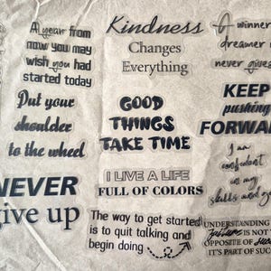 May include: A collection of clear stickers with motivational quotes in a variety of fonts. Phrases include "Never give up," "Keep pushing forward," and "I live a life full of colors." The stickers are on a textured, off-white background.