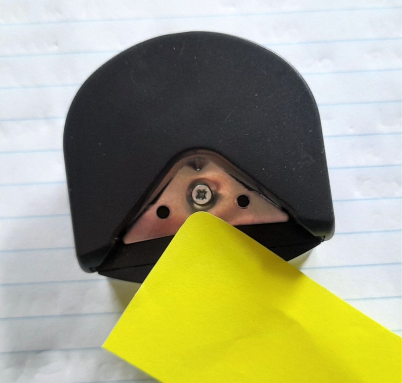 May include: A black corner rounder tool with a metal cutting edge. A yellow piece of paper is being placed into the corner rounder.