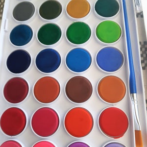 May include: A white plastic watercolor paint set with 24 round paint pans. The pans are arranged in a grid pattern and contain various colors, including black, brown, green, blue, orange, red, and pink. A blue and white paintbrush is included.