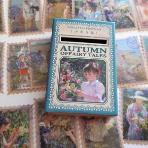 May include: A teal blue box with a slot on the top. The box has a picture of a girl in a pink dress holding a flower. The text on the box reads "The Little Postman (少女与花) Autumn Offairy Tales".