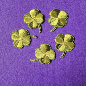 May include: Five embroidered four-leaf clover patches in gold and green sit on a purple background. Each clover has detailed stitching and a green stem, suitable for crafts or embellishing clothing.