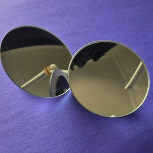 Può includere: Due specchi rotondi di colore argento su una superficie viola. Gli specchi sono leggermente inclinati l'uno verso l'altro, riflettendo l'ambiente circostante.