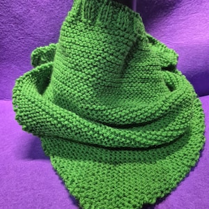 Puede incluir: Bufanda tejida a mano, de color verde brillante, colocada sobre una cabeza de maniquí negra. La bufanda tiene forma triangular y presenta un patrón de tejido texturizado. El color verde vibrante contrasta con el fondo morado, resaltando la calidad artesanal.