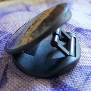 May include: A black phone grip with a circular mirror on top. The grip has a foldable design and a textured surface.