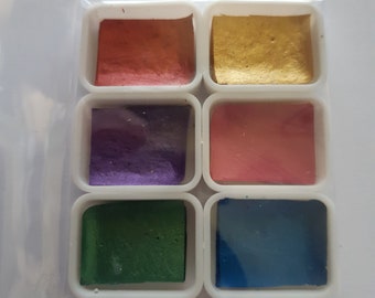 Puede incluir: Un juego de seis pastillas de pintura acuarela en un estuche de plástico. Las pastillas están dispuestas en dos filas de tres. Los colores son rojo, dorado, morado, rosa, verde y azul.