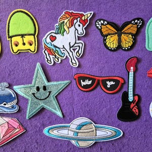 May include: A collection of embroidered iron-on patches featuring a yellow pineapple, a green bear head, a white unicorn, a monarch butterfly, a green elephant, a pink donut, a blue whale, a glittery star, red sunglasses with text, a guitar, lips, a diamond, a planet, and a hot air balloon.