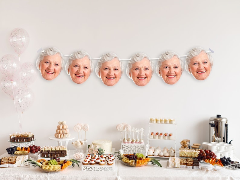 May include: A white table with a white tablecloth and a variety of desserts and snacks. There is a white banner with photos of older women's faces hanging above the table.