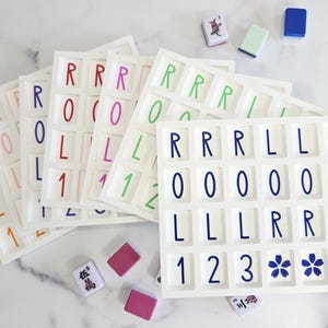 May include: White game boards with colorful letters and numbers, along with game tiles. The boards spell out "ROLL 1" and "ROLL 0" in various colors. Additional tiles are scattered around the boards, including some with floral designs.