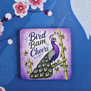 May include: Square coaster with a purple background featuring a peacock perched on bamboo. The peacock has a purple head and neck, green and blue body, and a large, detailed tail. The text "Bird Bam Cheers" is written in a decorative font.