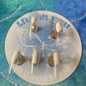 以下が含まれることがあります： 白い円形のディスプレイに、白い先端のついた透明な付け爪が6本。ディスプレイには、薄いグレーのフォントで「これまでの人生をありがとう」という文字が書かれている。