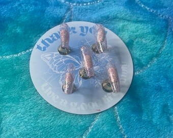 Peut inclure: Un ensemble de six faux ongles transparents avec des paillettes roses et argentées. Les ongles sont présentés sur un cercle blanc avec le texte "thank you" et "live a good life" dessus.