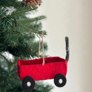 May include: A red felt wagon ornament featuring a black handle and four black wheels with white centers. The wagon is suspended by a thin brown twine string, designed for hanging on a tree branch as a decorative holiday ornament.