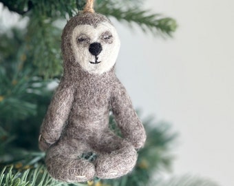 May include: A gray felt sloth ornament with a white face and closed eyes, hanging from a brown twine string on a green Christmas tree.