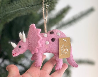 May include: Pink felt triceratops ornament with white and brown spots. The ornament is hanging from a brown twine string. The ornament has a brown paper tag with the text "ganapati crafts uk" on it.