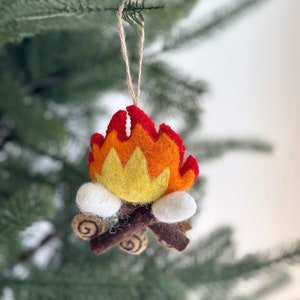 Puede incluir: Adorno de Navidad de fieltro con forma de fogata con llamas rojas, naranjas y amarillas, troncos marrones y malvaviscos blancos.