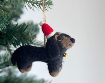 May include: A black and brown felt dachshund wearing a red and white Santa hat with a gold bell hanging from its neck. The dachshund is hanging from a string and is a Christmas ornament.