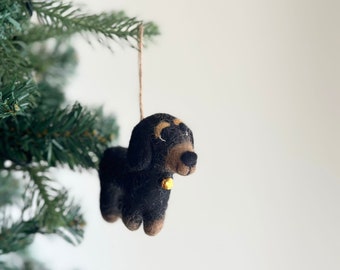 May include: A black and brown felt dachshund ornament with a gold bell hanging from a brown string. The ornament is hanging from a Christmas tree.