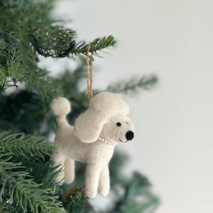 Könnte beinhalten: Weißer Filz-Pudel-Weihnachtsbaumschmuck mit einer Perlenkette. Der Anhänger hängt an einer braunen Schnur an einem grünen Weihnachtsbaum.