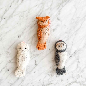 May include: Three felt owl ornaments, one brown, one white, and one gray. The owls are all stitched with black thread and have black eyes.