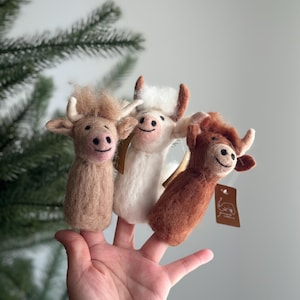 May include: Three felt finger puppets shaped like cows. The cows are brown, white, and tan. The puppets are all smiling and have black eyes. The puppets are on a person's hand.