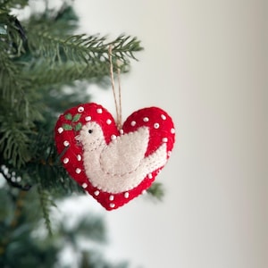 May include: A red felt heart ornament with a white felt dove and green felt leaves inside. The heart is decorated with white beads.