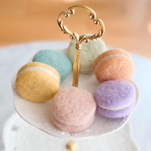 Peut inclure: Six macarons en feutre de couleur pastel sur un présentoir à gâteaux à étages blanc avec une poignée en métal doré. Les macarons sont disposés en cercle autour de la poignée.