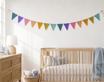 May include: A decorative bunting banner featuring a series of triangular fabric flags in various colors including teal, purple, pink, orange, yellow, and green, suspended horizontally by a thin string against a plain white wall surface.