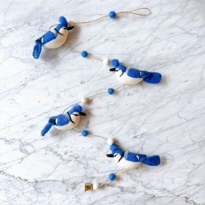 May include: A garland featuring four blue jay birds with white bellies, strung with white and blue pom poms and twine. The garland is hanging on a white marble surface.