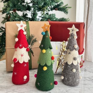 May include: Three cone-shaped felt Christmas tree decorations in red, green, and gray. Each tree features scalloped layers, small white and colored pom-poms, embroidered star patterns, and a felt star topper. The red tree is white-accented, the green tree has a yellow star, and the gray tree features white and gray details.