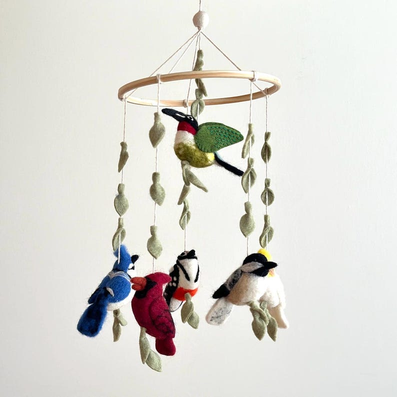 Puede incluir: Un m&oacute;vil de madera con cinco p&aacute;jaros de fieltro y hojas verdes de fieltro colgando de un marco circular de madera. Los p&aacute;jaros incluyen un colibr&iacute;, un arrendajo azul, un cardenal, un p&aacute;jaro carpintero velloso y un carbonero.