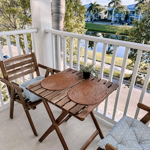 May include: A wooden table with two chairs on a balcony overlooking a pond and a residential area. The table has two brown placemats and a small potted plant. The chairs have blue and white patterned cushions.