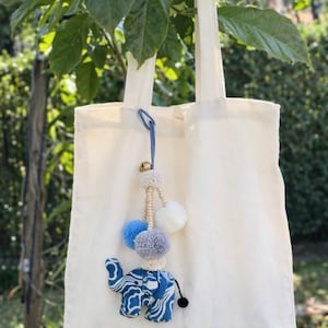 Può includere: Una borsa tote color crema con un ciondolo a forma di elefante con motivi blu e bianchi. Il ciondolo include una campana, pompon bianchi, blu e grigi e un cordino color crema. La borsa è appesa a un ramo d'albero.