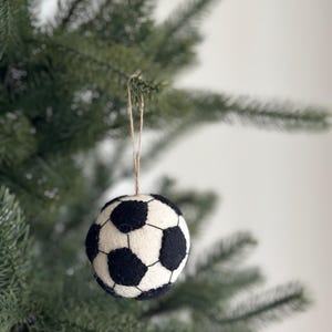 May include: A soccer ball ornament, featuring a classic black and white design, hangs from a natural twine string. The ornament is suspended against a backdrop of green Christmas tree branches, creating a festive holiday scene.