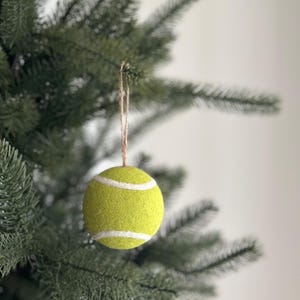 May include: A tennis ball Christmas tree ornament, bright yellow-green with white stripes, suspended by twine. The decoration hangs on a green Christmas tree, offering a sporty and festive holiday theme. The ball's diameter is approximately 6.7 cm.
