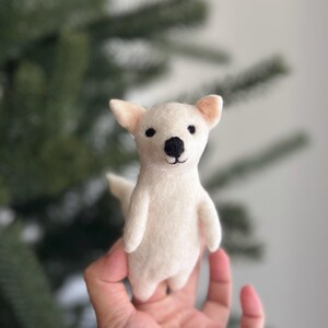 May include: A small, white, felt dog toy with black eyes and a black nose. The toy is being held in a person's hand.