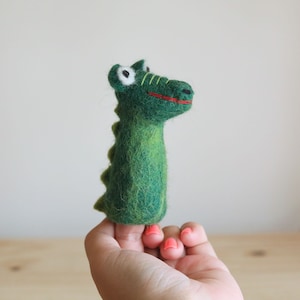 May include: Green felt finger puppet shaped like a dinosaur with white eyes and a red mouth.