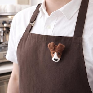Può includere: Un grembiule marrone con una spilla a forma di testa di cane in feltro. La spilla a forma di cane ha pelo marrone, segni bianchi e un naso nero. Il grembiule è indossato sopra una camicia bianca.