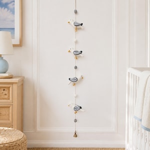 Pode incluir: Um móbile vertical suspenso com quatro figuras de gaivota em feltro com corpos brancos e cinzentos e patas amarelas, intercaladas com pequenas bolas de feltro cinzentas e brancas num fio fino, terminado com um pequeno sino metálico na ponta.