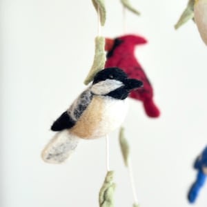 Puede incluir: Un m&oacute;vil de p&aacute;jaros de fieltro con un carbonero blanco y negro, un cardenal rojo y hojas verdes.