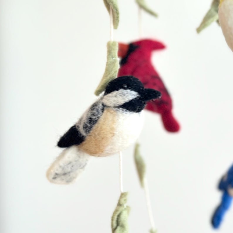Puede incluir: Un m&oacute;vil de p&aacute;jaros de fieltro con un carbonero blanco y negro, un cardenal rojo y hojas verdes.