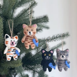 Könnte beinhalten: Vier Filzkatzen-Anhänger, die an einem Weihnachtsbaum hängen. Jede Katze hält einen blauen Filzfisch. Die Katzen sind braun-weiß gestreift, weiß-orange gestreift, schwarz und grau.