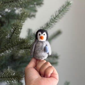 May include: A handmade felt penguin ornament with a gray body, black head, white face, and orange beak. The penguin holds a small blue object. The ornament is displayed in front of a green Christmas tree.