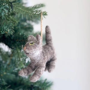 May include: A gray felt cat ornament with green eyes hanging from a brown string on a green Christmas tree.