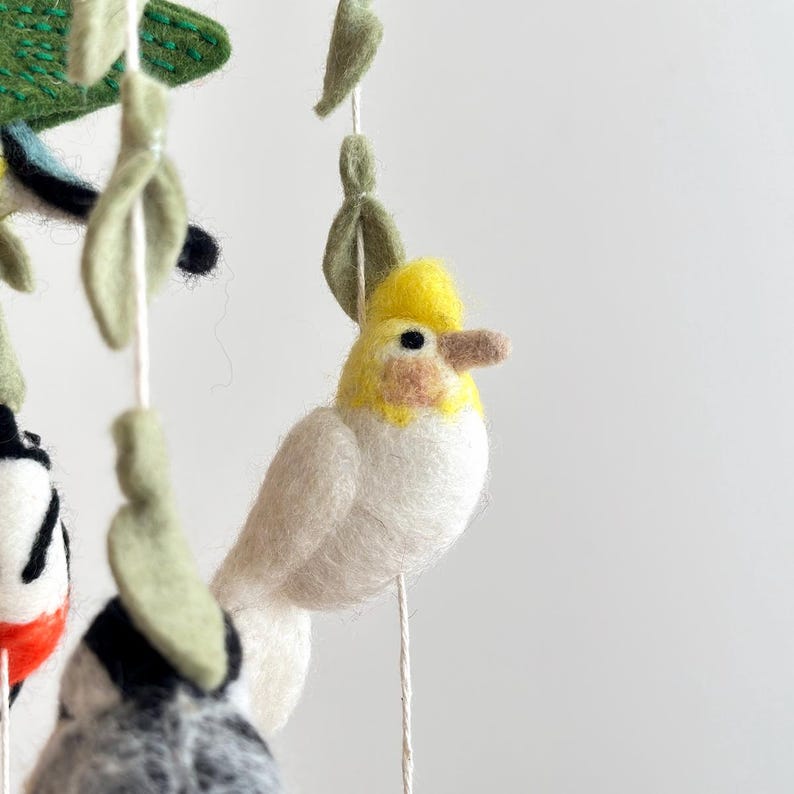 Puede incluir: Un p&aacute;jaro de fieltro blanco y amarillo con una cresta amarilla que cuelga de un m&oacute;vil. El p&aacute;jaro tiene un cuerpo blanco y una cabeza amarilla con un ojo negro. El p&aacute;jaro est&aacute; colgado de una cuerda blanca.