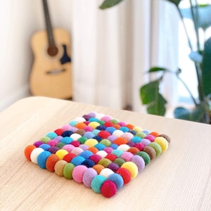 May include: A square, multi-colored felt ball trivet. The trivet is made of numerous small, round felt balls in various colors, including red, orange, yellow, green, blue, purple, and white. The trivet sits on a light-colored wooden surface.